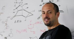 A man with a short beard and mustache standing in front of a whiteboard covered with chemical equations and diagrams.