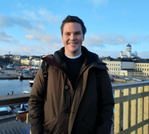 Man in a brown jacket smiling at the camera. Water and buildings in the background.